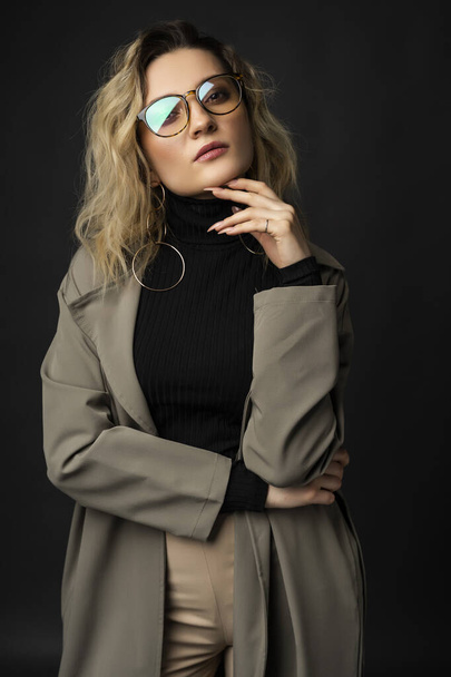 Beautiful girl wearing a casual trendy trousers, turtleneck glasses and raincoat posing on a dark background. Fashionable, advertising, lifestyle and commercial design - Photo, Image