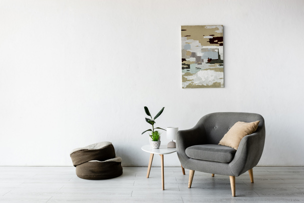 άνετη πολυθρόνα και μαξιλάρι κοντά στο τραπέζι του καφέ με πράσινα φυτά, λάμπα και ζωγραφική στον τοίχο στο σαλόνι  - Φωτογραφία, εικόνα