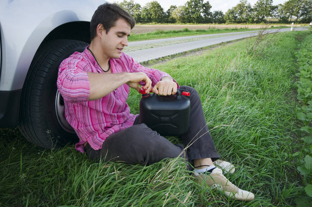 Vista lateral de corpo inteiro de um jovem sentado na beira de uma estrada rural na grama no pneu dianteiro de seu carro abrindo o tampão da lata
. - Foto, Imagem
