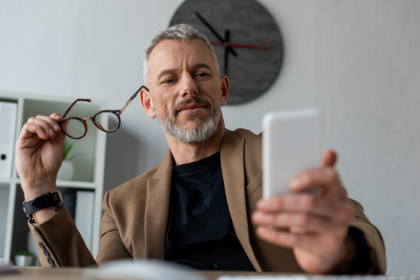 επιλεκτική εστίαση του επιχειρηματία κρατώντας γυαλιά και κοιτάζοντας smartphone στο γραφείο  - Φωτογραφία, εικόνα