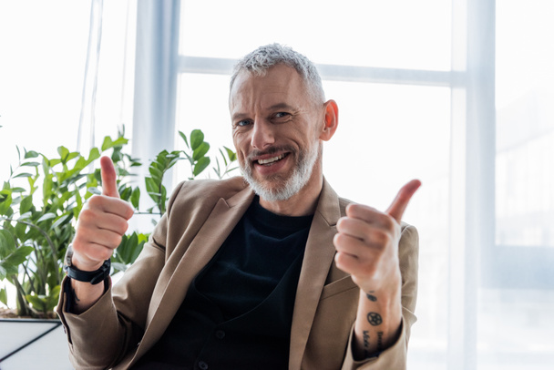 happy and tattooed businessman showing thumbs up while looking at camera  - Photo, Image
