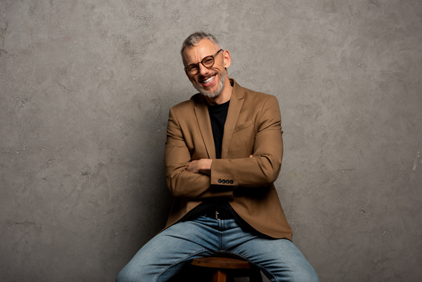 hombre de negocios barbudo en gafas mirando a la cámara y sentado con los brazos cruzados en gris
  - Foto, imagen