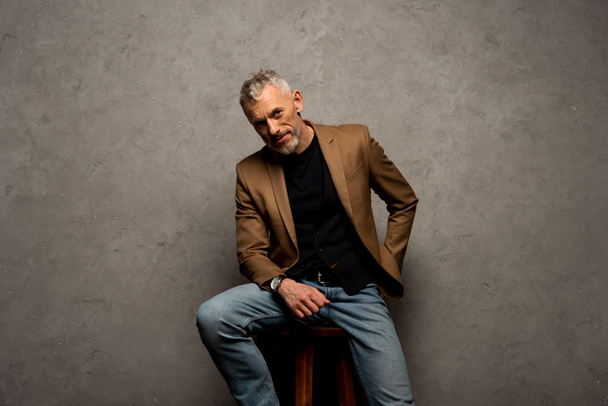 businessman in jeans and blazer sitting on stool and looking at camera on grey  - Foto, afbeelding