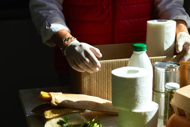 Εθελοντής στην προστατευτική ιατρική μάσκα και γάντια βάζοντας φαγητό σε κουτί δωρεάς. Υπάλληλος παράδοσης σε κόκκινο γιλέκο συσκευασίας κουτί με φαγητό. Υπηρεσία καραντίνας πανδημία coronavirus. - Φωτογραφία, εικόνα