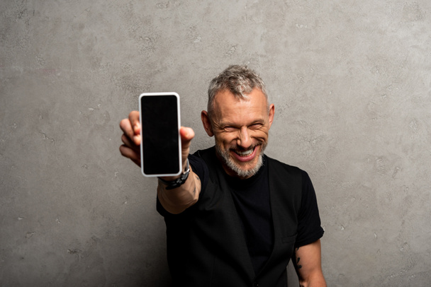 幸せな男の選択的な焦点は灰色の上に空白の画面を持つスマートフォンを保持  - 写真・画像