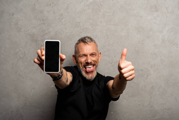 szelektív fókusz boldog ember kiálló nyelv és gazdaság okostelefon üres képernyőn, miközben mutatja hüvelykujját fel szürke  - Fotó, kép