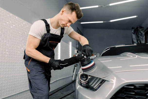 Car detailing and polishing concept. Professional Caucasian male car service worker, holding in hands orbital polisher, and polishing white luxuty car in auto repair shop. - Zdjęcie, obraz