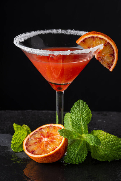 Primer plano de copa de cóctel con martini naranja sangre y azúcar, con media naranja y menta, sobre fondo negro, en vertical
 - Foto, imagen