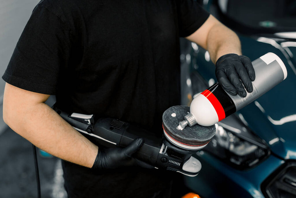 Cropped image of hands of male auto service worker, putting special polish wax or cream on the orbital polish machine to polish the blue car on the background. Car detailing and polishing concept - Fotoğraf, Görsel