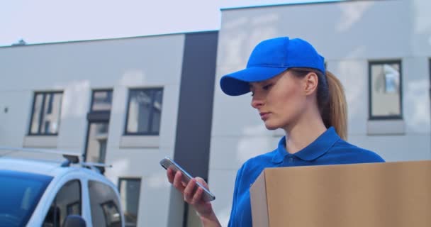 Close up of Caucasian young pretty woman in blue uniform and hat holding carton box and tapping or texting message on smartphone outdoor. Attractive female courier typing and scrolling on phone. - Imágenes, Vídeo
