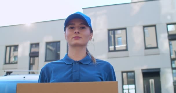 Close up of young Caucasian beautiful woman in blue uniform and hat coming to camera with parcel in hands. Pretty female courier handing parcel to camera and smiling. - Záběry, video