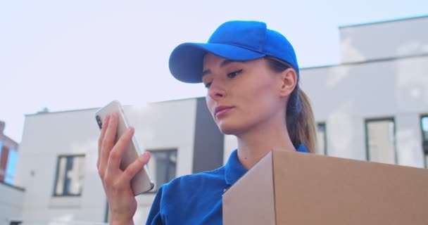 Beautiful young Caucasian female delivery worker in blue cap and with carton box tapping on smartphone outdoors. Pretty girl texting sms and scrolling on mobile phone at street. Delivering concept. - Video