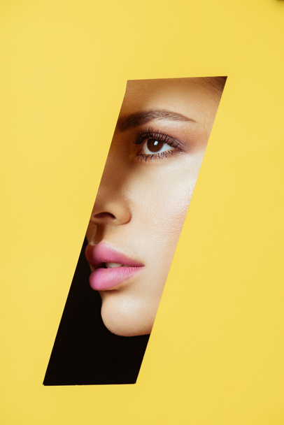 Female face with makeup in quadrangular hole in yellow paper on black - Photo, Image