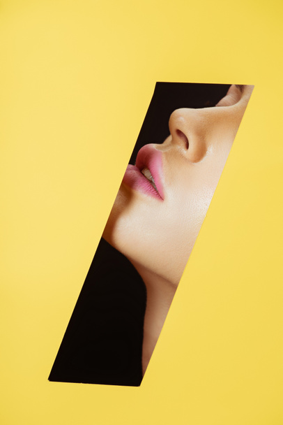 Vue recadrée du visage féminin avec lèvres roses dans un trou quadrilatéral en papier jaune sur fond noir
 - Photo, image