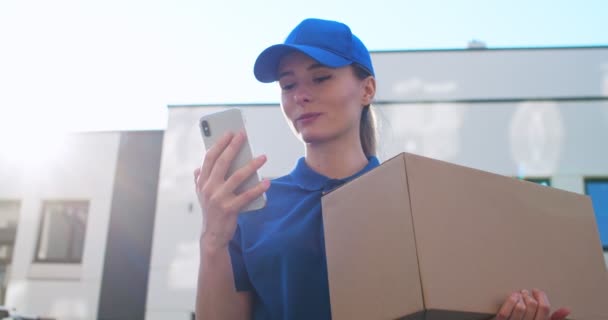 View from below on beautiful young Caucasian female postal worker in blue cap and with carton box tapping on smartphone outdoors. Pretty girl texting sms and scrolling on mobile phone at street. - Materiaali, video