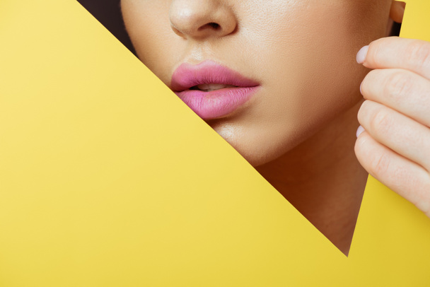 Cropped view of woman with pink lips in triangular hole touching yellow paper on black - Fotografie, Obrázek