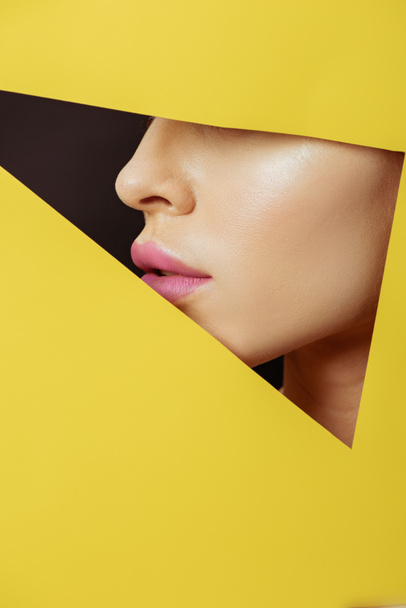 Cropped view of female face with pink lips in triangular hole in yellow paper on black - Fotografie, Obrázek
