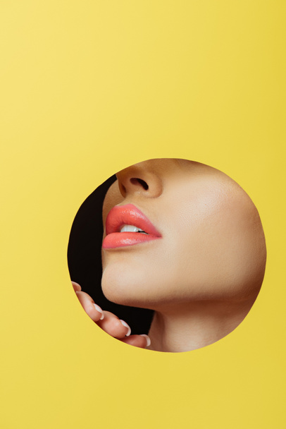 Cropped view of female face with coral lips and hand in round hole in yellow paper on black - Foto, Bild