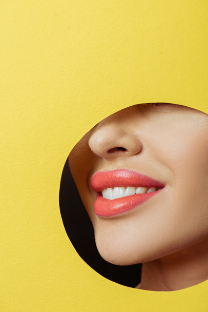 Cropped view of woman with coral lips smiling in round hole in yellow paper on black background - Foto, Imagem