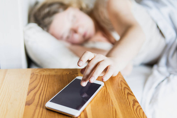 Young Eastern European woman lying in bed and turning off the alarm clock on her mobile phone in the morning. - Photo, image