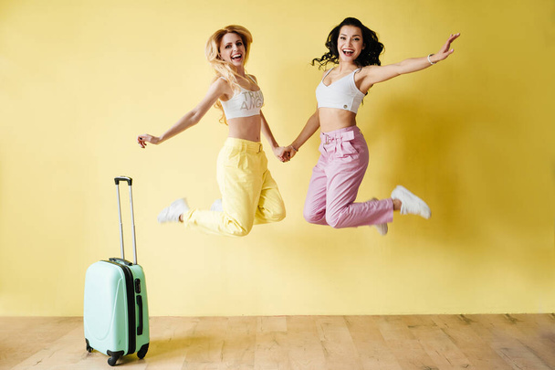Brunette and blonde in colorful trousers on a yellow background. Pink and yellow colour. Traveling with a mint suitcase and pinapple. - Photo, image