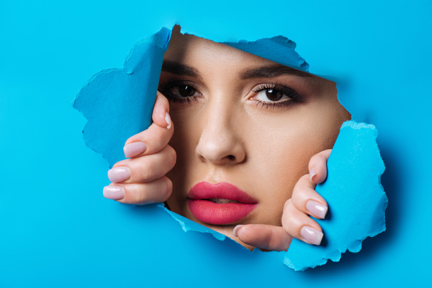 Beautiful woman with smoky eyes and pink lips looking at camera across hole and touching blue paper - Фото, зображення