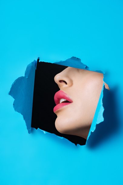 Female face with pink lips in hole in blue paper on black - Foto, immagini