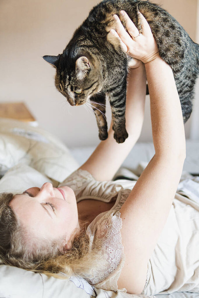 Młoda i uśmiechnięta wschodnioeuropejska kobieta leżąca rano w łóżku z czarnym kotem w rękach. - Zdjęcie, obraz