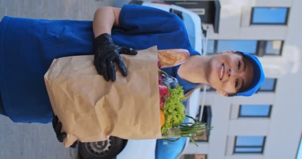 Vertical portrait of beautiful young woman of delivery service, in blue uniform, gloves and cap smiling to camera outdoor with fresh food in packet. Pretty female courier of grocery. Stories concept. - Filmagem, Vídeo