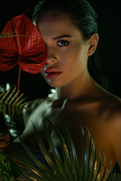 Girl looking at camera and covering face with red anthurium near leaves isolated on black - Foto, immagini