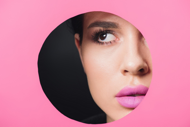 Beautiful girl with smoky eyes and pink lips looking away across round hole in paper on black  - Photo, Image