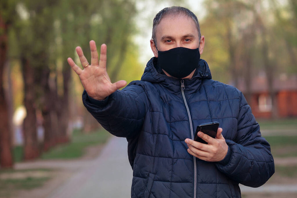 Młody facet w czarnej masce na twarzy i z telefonem w ręku, patrzący w kamerę na zewnątrz, pokazujący dłoń, dłoń, nie zatrzymujący się. Zanieczyszczenie powietrza, wirus, koncepcja chińskiego pandemicznego koronawirusu. - Zdjęcie, obraz