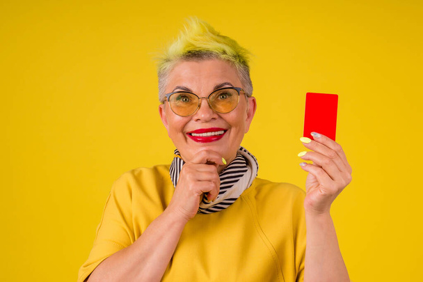 mature old woman with colored stylish yellow dyed hair and red lipstick lips in warm dress holding credit card background studio:black friday sale - Φωτογραφία, εικόνα