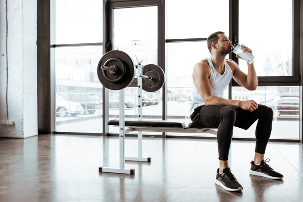 handsome sportsman drinking protein milkshake near barbell in gym  - Fotoğraf, Görsel
