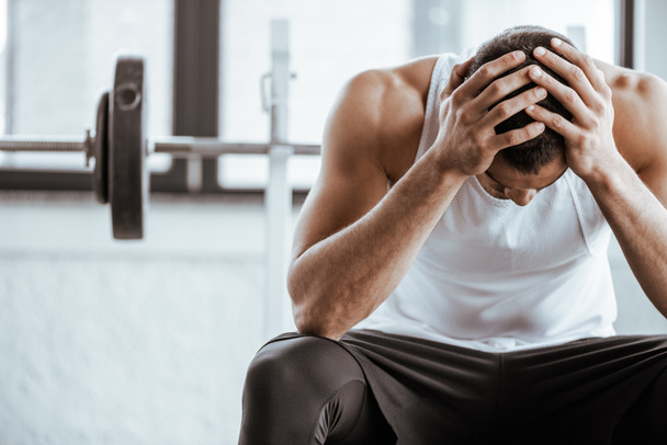 upset sportsman touching head while sitting near barbell in gym  - Фото, зображення