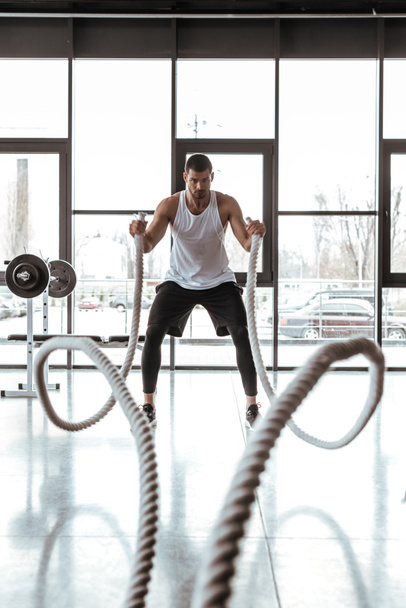 selective focus of concentrated sportsman exercising with battle ropes in modern gym  - Photo, Image