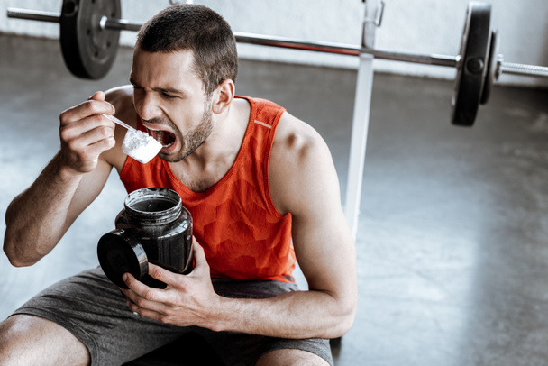 αθλητικός αθλητής με ανοιγμένο στόμα βαζάκι συγκράτησης και κουτάλι μέτρησης με σκόνη πρωτεΐνης  - Φωτογραφία, εικόνα