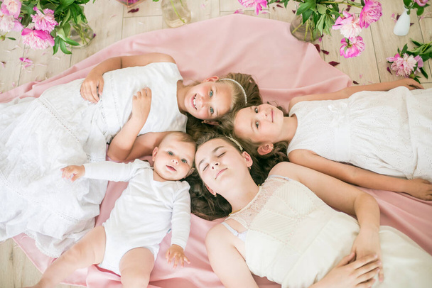 Милые дети, младший брат и старшие сестры в белых платьях лежат вместе на полу в яркой весенней студии с розовыми пионами. Счастливая семья. Счастливого детства
 - Фото, изображение