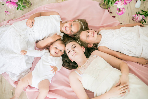 Niedliche Kinder, ein kleiner Bruder und ältere Schwestern in weißen Kleidern liegen zusammen auf dem Boden in einem hellen Frühlingsstudio mit rosa Pfingstrosen. Glückliche Familie. Glückliche Kindheit - Foto, Bild