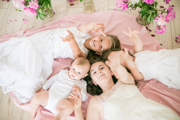 Słodkie dzieci, młodszy brat i starsze siostry w białych sukienkach leżą razem na podłodze w jasnym wiosennym studio z różowymi piwoniami. Szczęśliwa rodzina. Szczęśliwego dzieciństwa - Zdjęcie, obraz