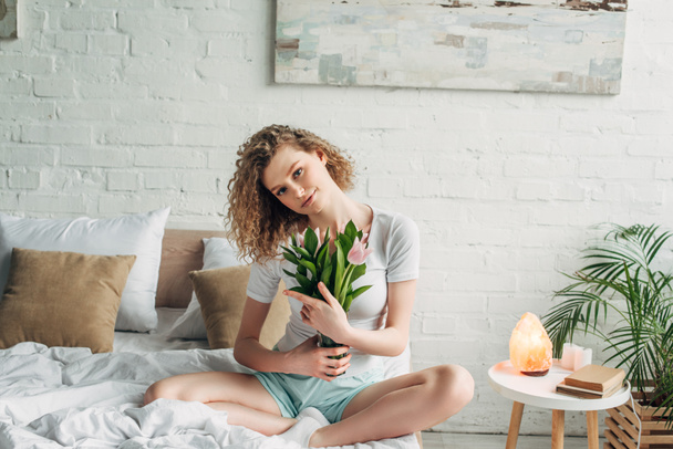 кудрявая девушка в домашней одежде держит тюльпаны в спальне с гималайской соляной лампой
 - Фото, изображение