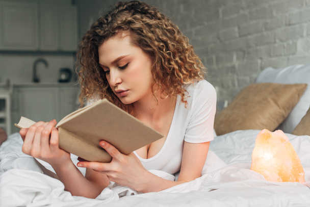 attractive curly girl reading book on bed with Himalayan salt lamp - Photo, Image