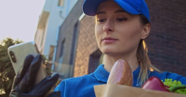 Close up of Caucasian young pretty woman in blue hat and gloves holding food packet and tapping or texting message on smartphone outdoor. Female grocery courier typing and scrolling on phone. - Footage, Video