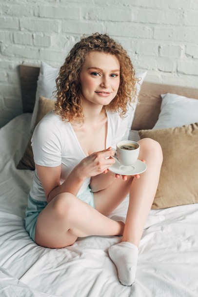 smiling curly girl in homewear holding cup of coffee on bed in morning  - Photo, Image