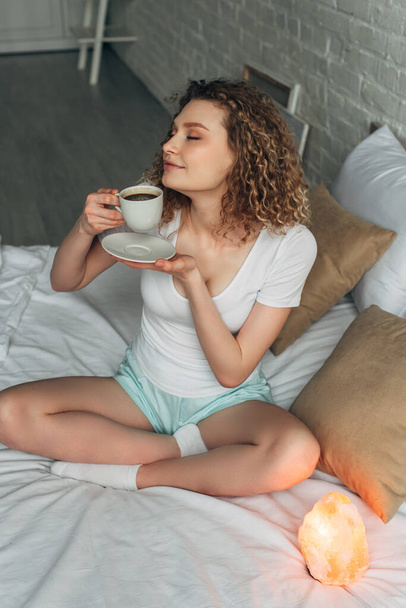 dreamy girl in homewear holding cup of coffee while sitting on bed with Himalayan salt lamp  - Foto, afbeelding