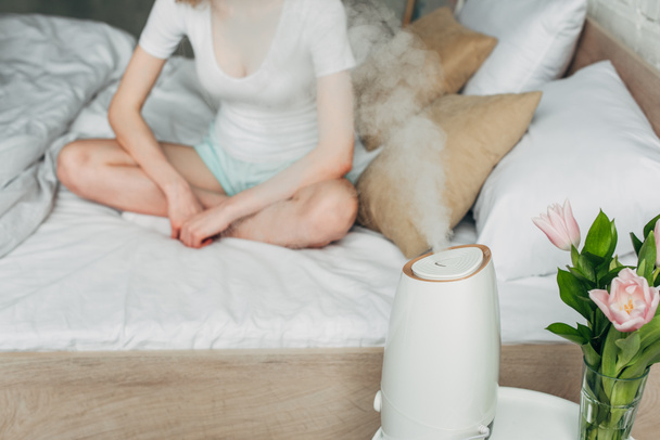 καλλιεργημένη άποψη του κοριτσιού σε homewear στο κρεβάτι με λουλούδια και καθαριστής αέρα εξάπλωση ατμού - Φωτογραφία, εικόνα