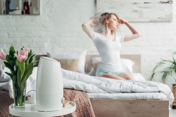 селективный фокус девушки, сидящей в спальне с цветами тюльпана и очистителем воздуха, распространяющим пар
 - Фото, изображение