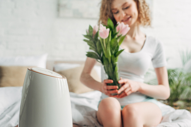 selective focus of smiling girl holding tulip flowers while sitting in bedroom with air purifier  - Photo, Image