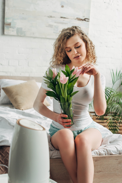 σγουρό κορίτσι κρατώντας λουλούδια τουλίπας, ενώ κάθεται στο υπνοδωμάτιο με καθαριστή αέρα  - Φωτογραφία, εικόνα