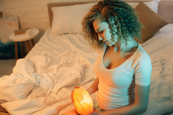 夜にベッドに座りながらヒマラヤの塩ランプを持っている美しい巻きガール - 写真・画像